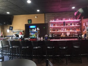 Zona de bar con diversas opciones de asientos, una barra con una variedad de botellas y una pantalla de televisión.