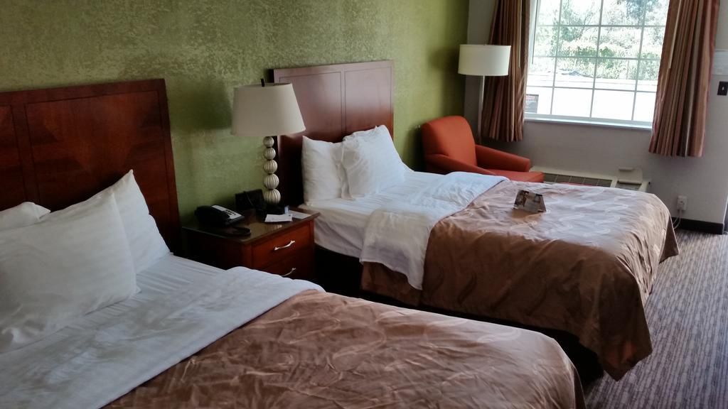 Habitación de hotel con dos camas, una mesita de noche con lámpara, una silla y una ventana con cortinas.