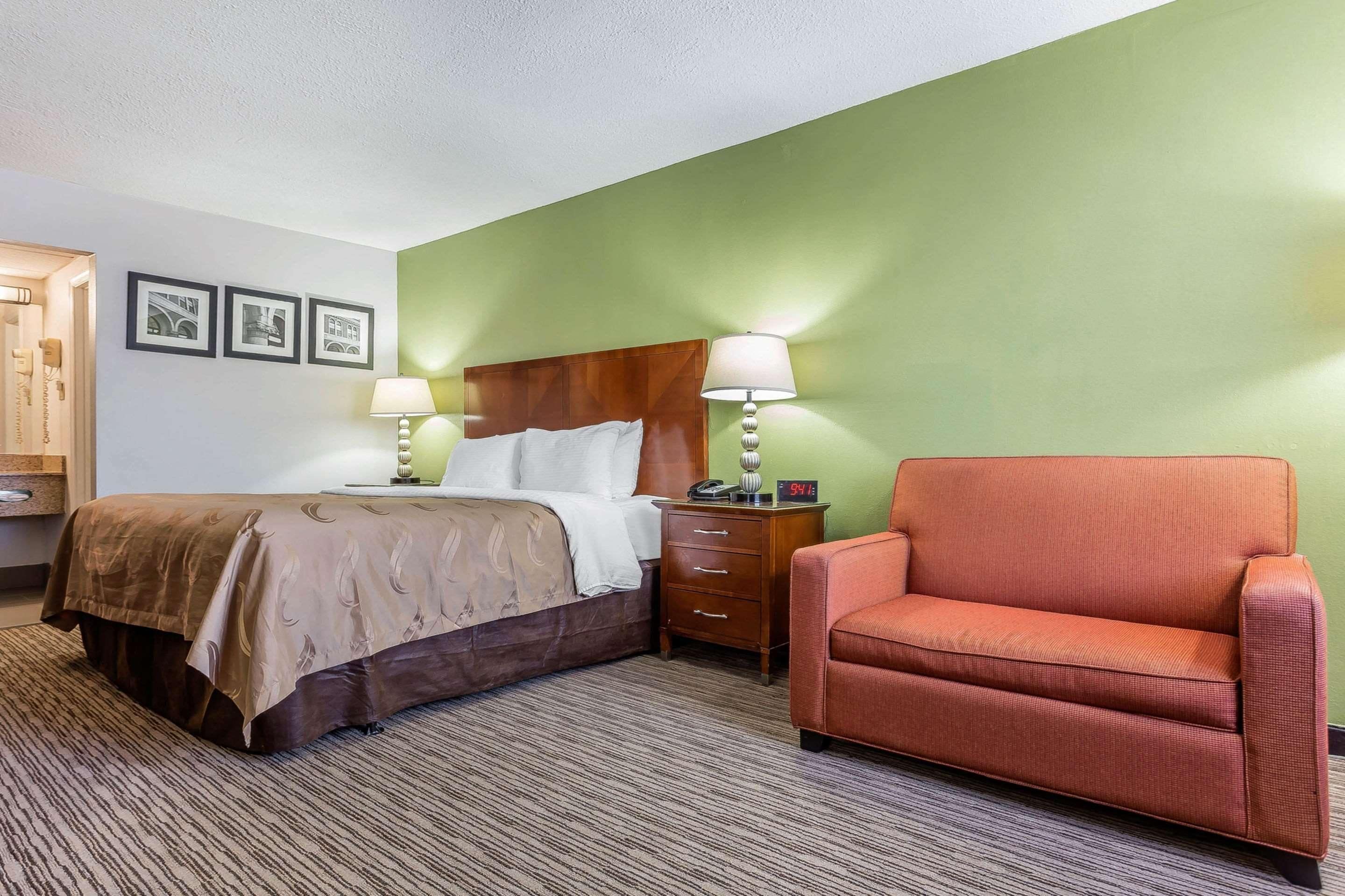 A hotel room with a bed, a nightstand, a chair, and a wall with framed pictures.