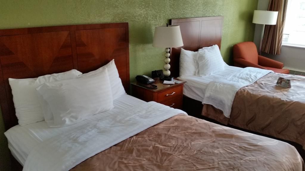 A hotel room with two beds, a nightstand with a lamp, and a chair.