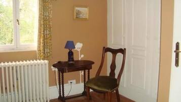 Una habitación con un radiador, una silla de madera, una mesita con una lámpara, un cuadro en la pared y una ventana con cortinas.