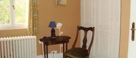 Una habitación con un radiador, una silla de madera, una mesita con una lámpara, un cuadro en la pared y una ventana con cortinas.