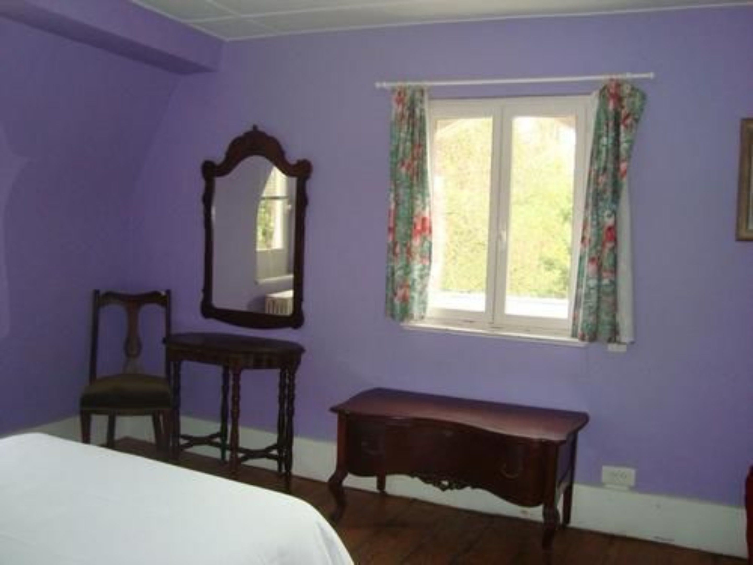 Une pièce comprenant un lit, une chaise, une table, une fenêtre avec des rideaux et un miroir au mur.