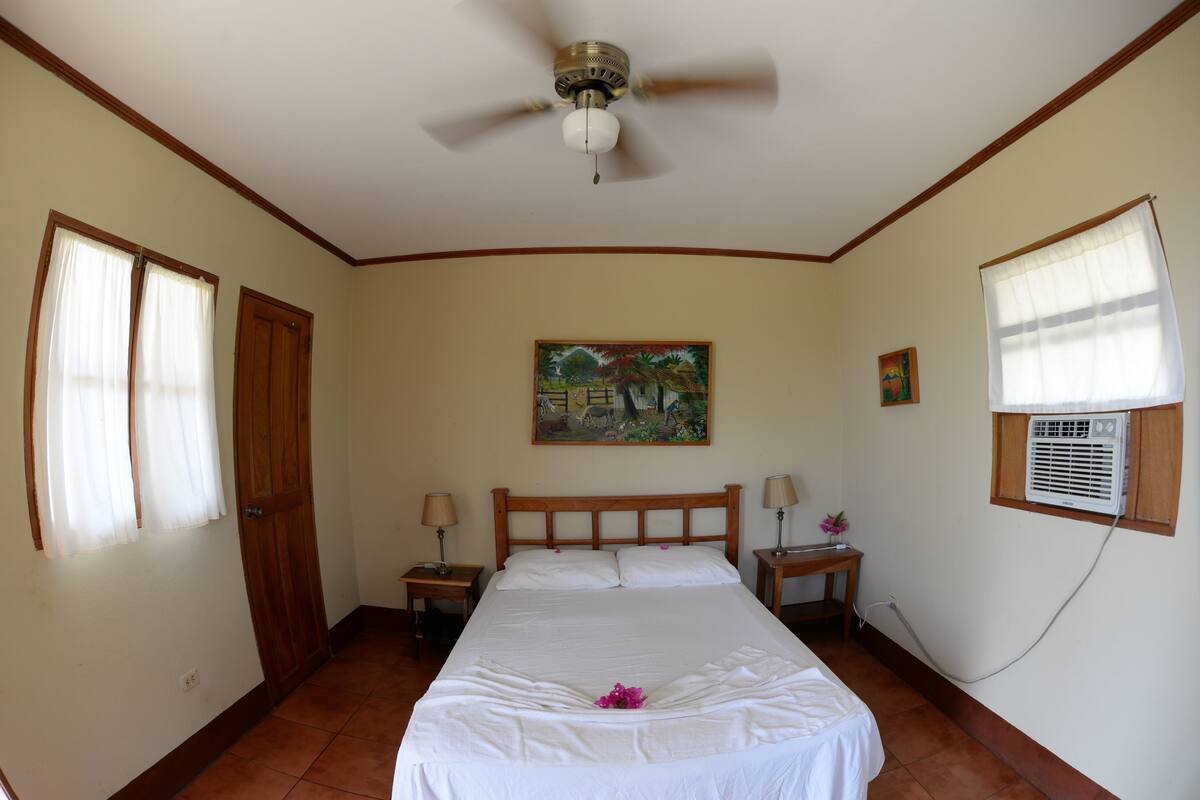 A bedroom with a bed, two bedside tables, a ceiling fan, a window with curtains, a wall-mounted air conditioner, and a painting on the wall.