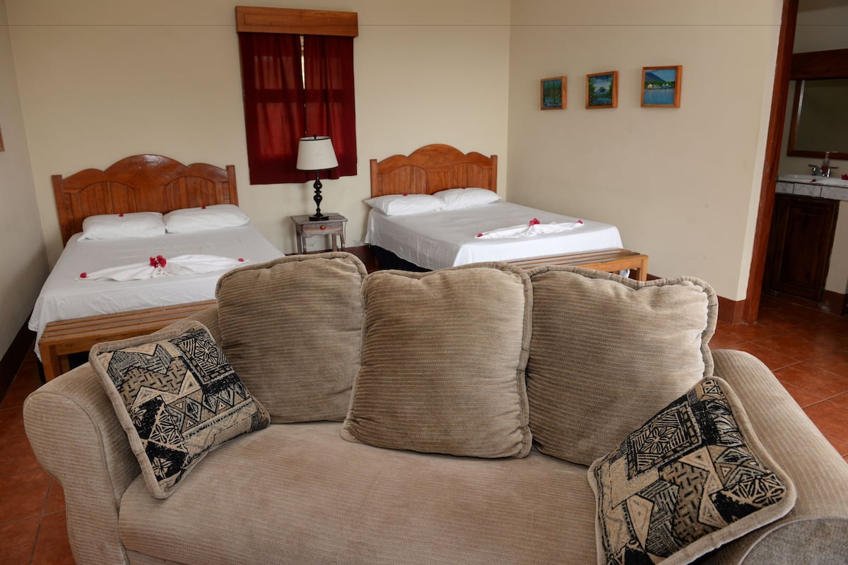 A hotel room with two beds, a wooden headboard, a small table, a lamp, and a couch with patterned cushions.