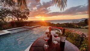 Une vue sur le coucher de soleil depuis un espace aménagé près de la piscine, avec une bouteille de vin et des verres sur une table.
