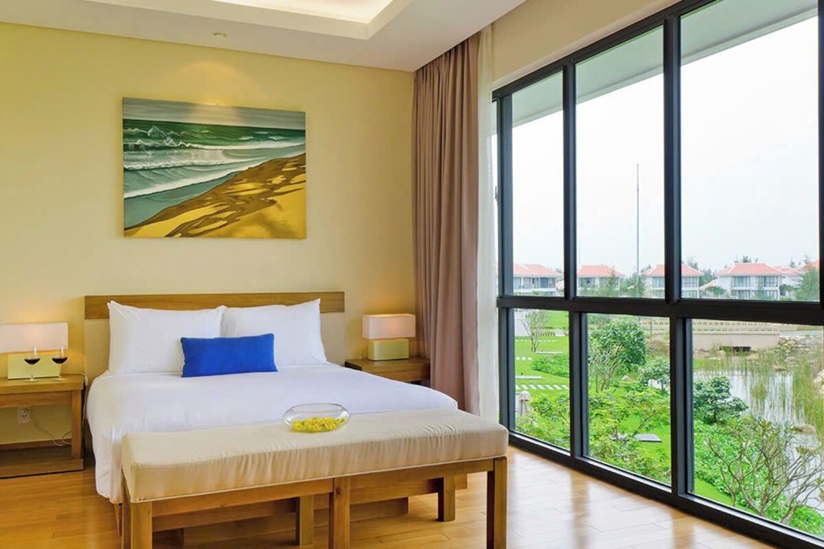 Een hotelkamer met een groot raam, een bed met witte lakens, een blauw kussen en een schilderij van een strand.