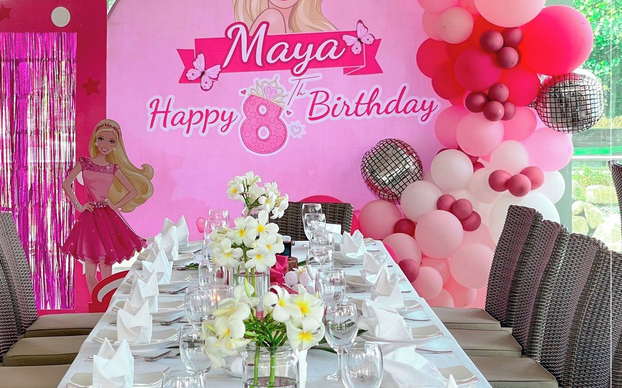 Une décoration pour une fête d’anniversaire avec une table dressée pour six personnes, un fond rose orné de l’inscription « 8e anniversaire de Maya » et des ballons.