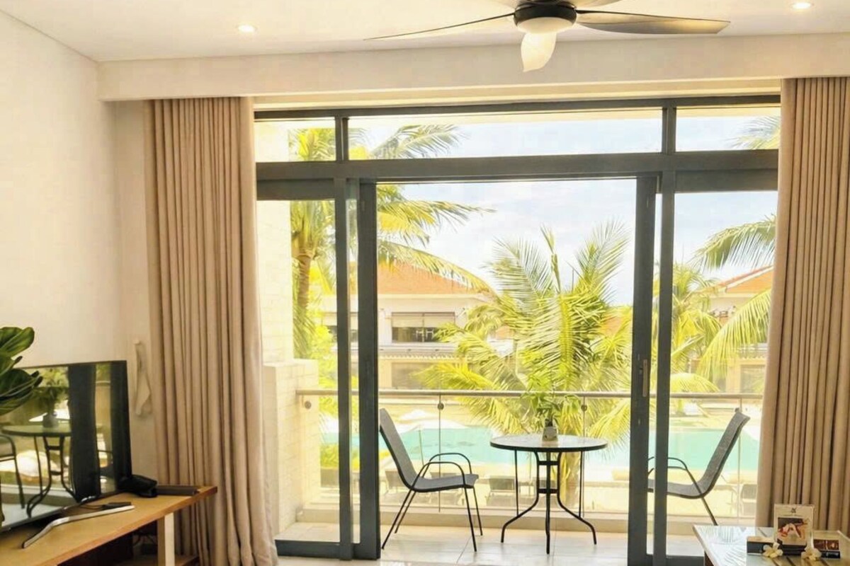 Hotelkamer met een bed, bureau, stoel, tafel, plafondventilator en groot raam met uitzicht op palmbomen en een zwembad.
