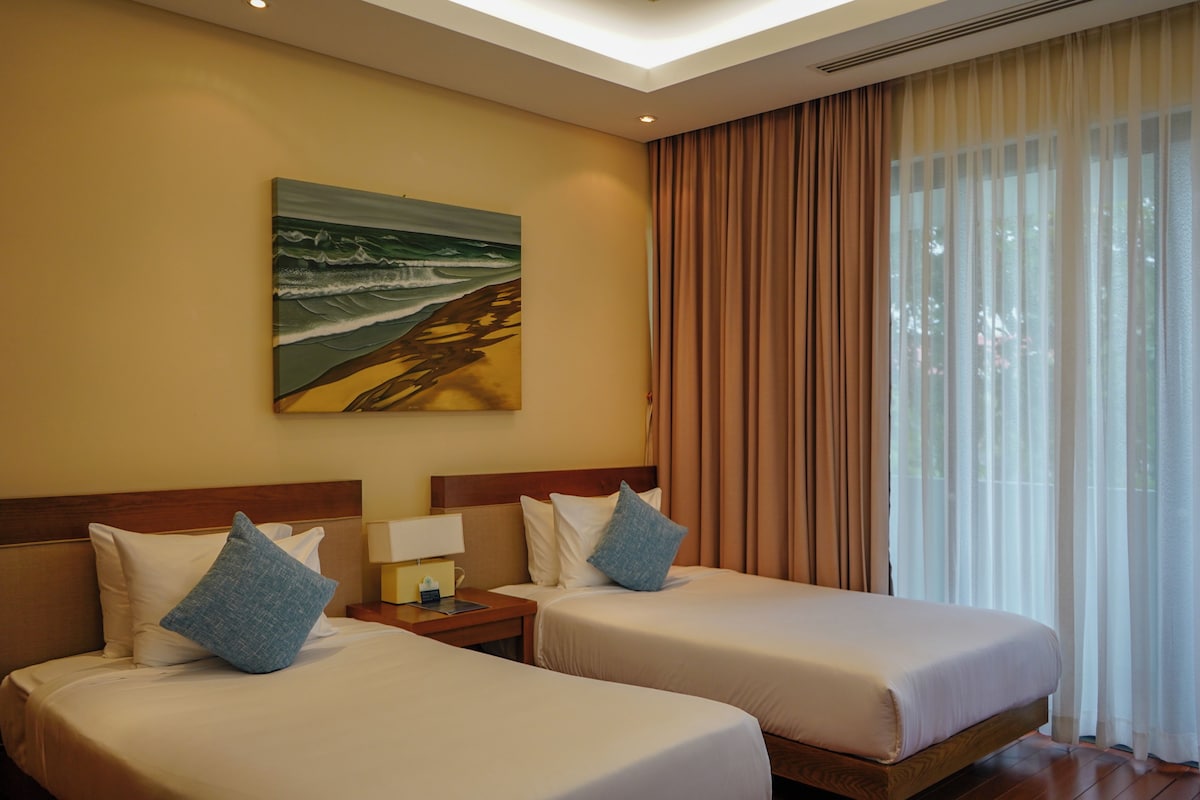 A hotel room with two beds, a painting of a beach scene, and a large window with curtains.