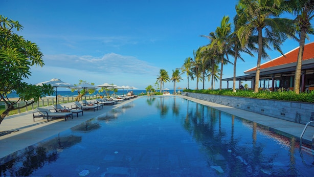 A swimming pool with lounge chairs and umbrellas, palm trees, and a view of the ocean. - The Ocean Villas (Da Nang)