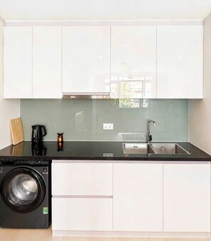 A compact kitchen with white cabinets, a stainless steel sink, a coffee maker, and a washing machine. - The Ocean Villas (Da Nang Municipality)