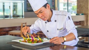 A chef in a white uniform with a tall hat, preparing a dish on a white plate. - The Ocean Villas (Da Nang Municipality)
