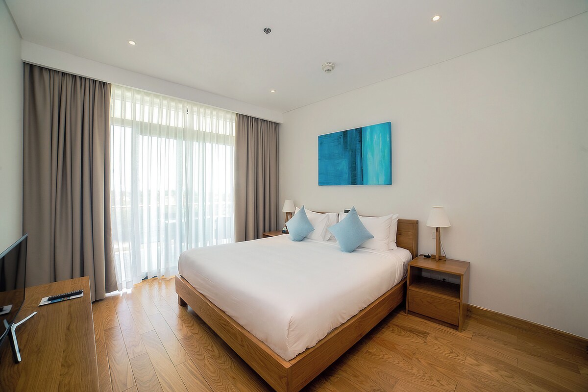 Een hotelkamer met een groot bed, een bureau, een stoel, een nachtkastje met een lamp en een schilderij aan de muur.