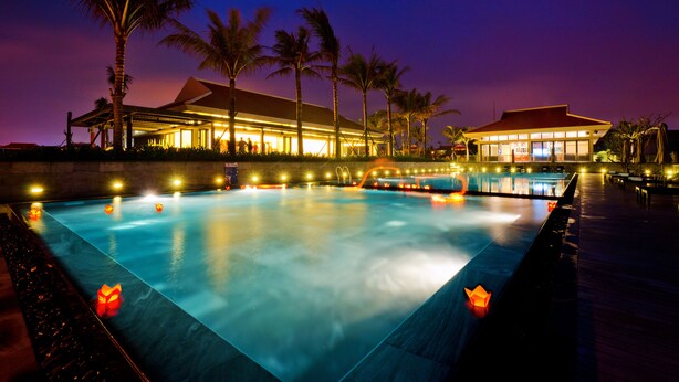 A large outdoor swimming pool with a wooden deck, surrounded by palm trees and a building with illuminated windows. - The Ocean Villas (Da Nang Municipality)