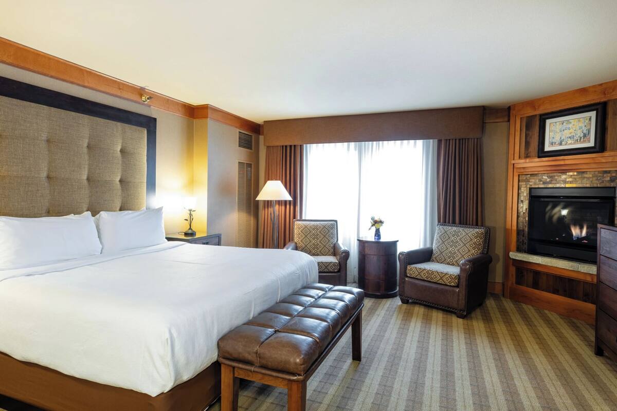 A hotel room with a large bed, a bench, two armchairs, a TV, and a fireplace.
