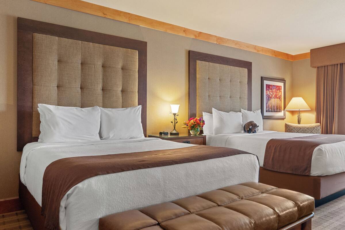 A hotel room with two beds, a brown leather bench, and a nightstand with a lamp and flowers.