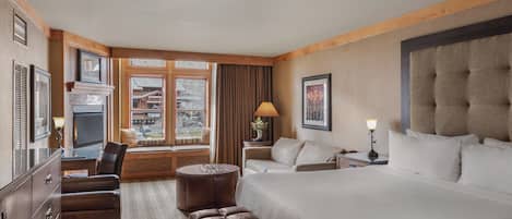 Habitación de hotel con una cama grande, un escritorio con silla, un sofá, un taburete redondo pequeño y una televisión, además de un ventanal con cortinas.