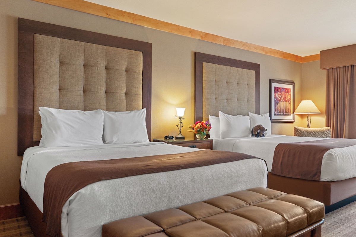 A hotel room with two beds, a brown leather bench, and a nightstand with a lamp and flowers.