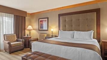 A hotel room with a large bed, a leather armchair, a wooden headboard, and a nightstand with a lamp.