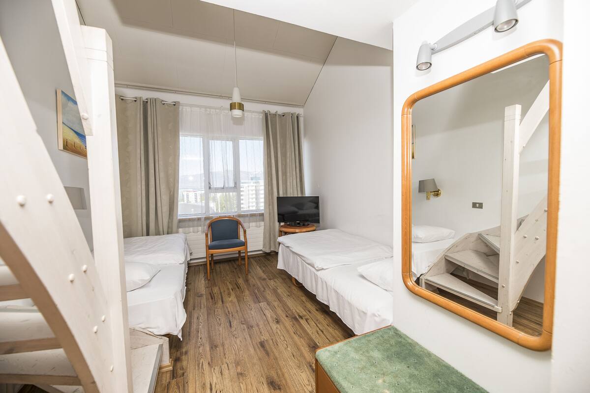 Ein Hotelzimmer mit zwei Einzelbetten, einem Fernseher, einem Sessel, einem Fenster mit Vorhängen und einem Spiegel.
