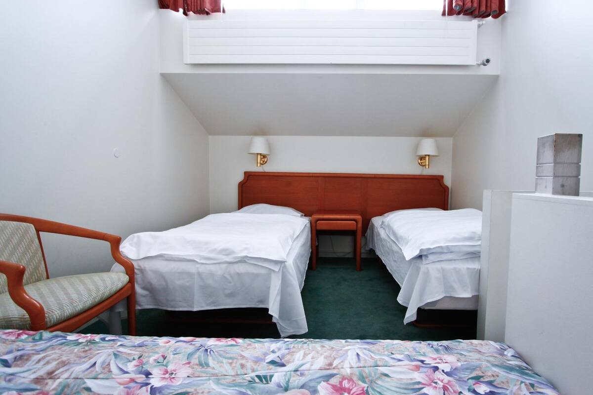 Ein kleines Hotelzimmer mit zwei Einzelbetten, einem Holzstuhl, einem Schreibtisch und einem Fenster mit Vorhängen.