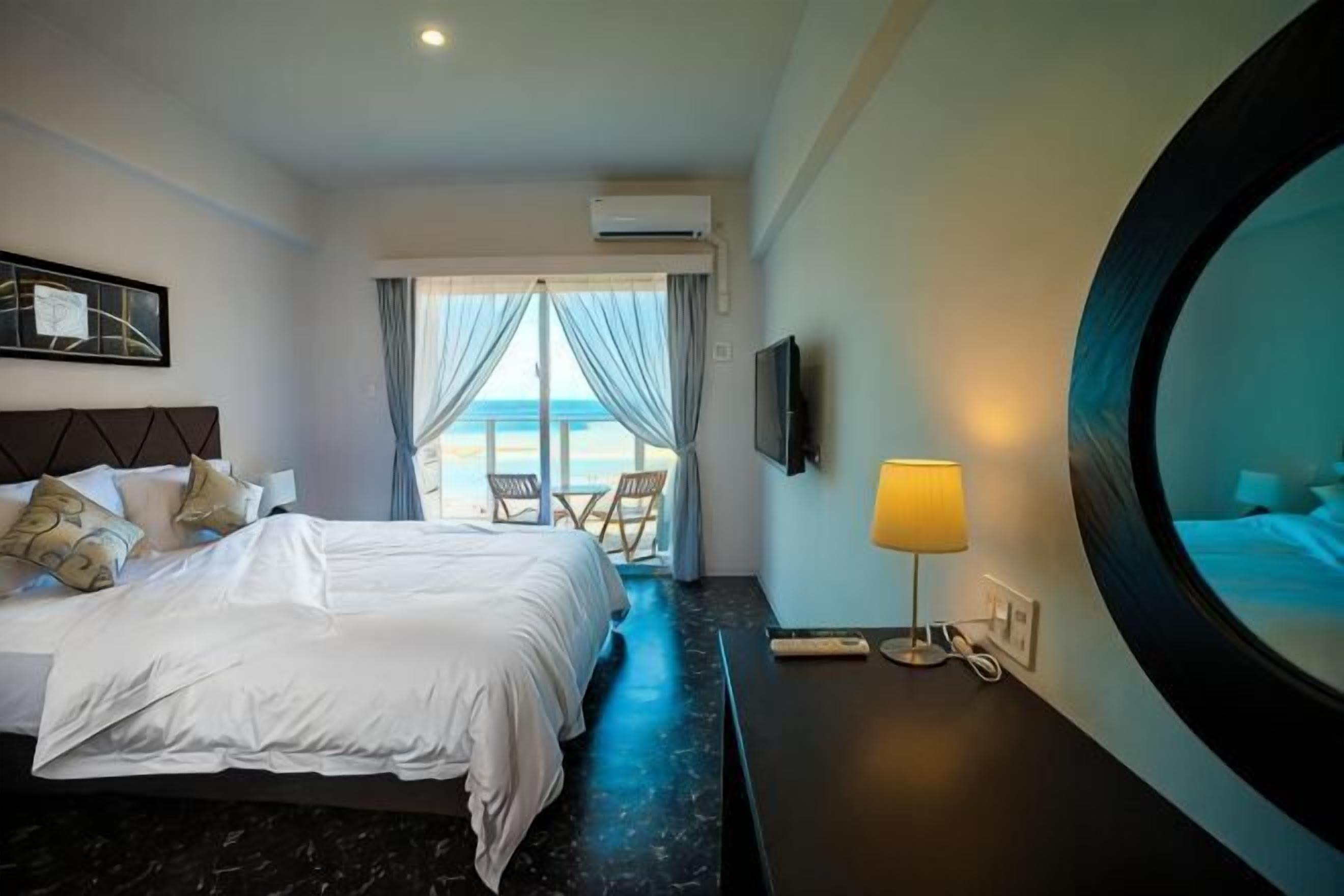 A hotel room with a bed, a TV, a lamp, a round mirror, and a view of the beach.