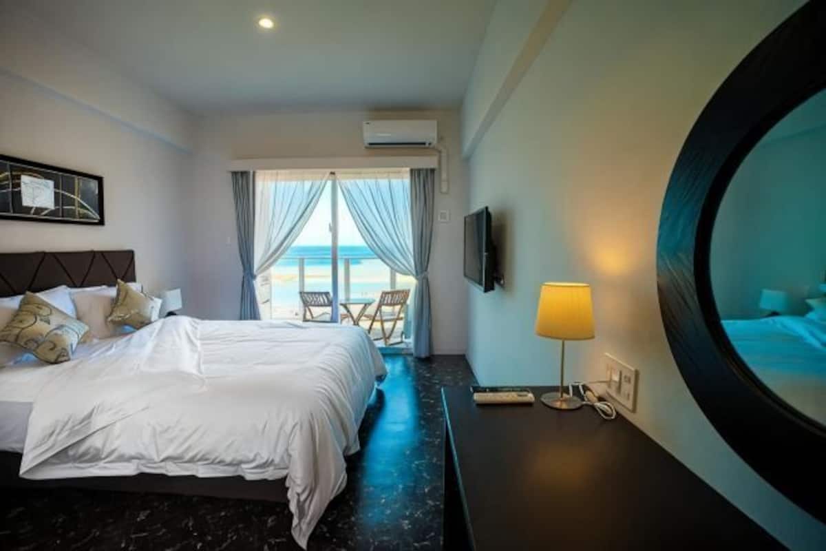 A hotel room with a bed, a TV, a lamp, a round mirror, and a view of the beach.