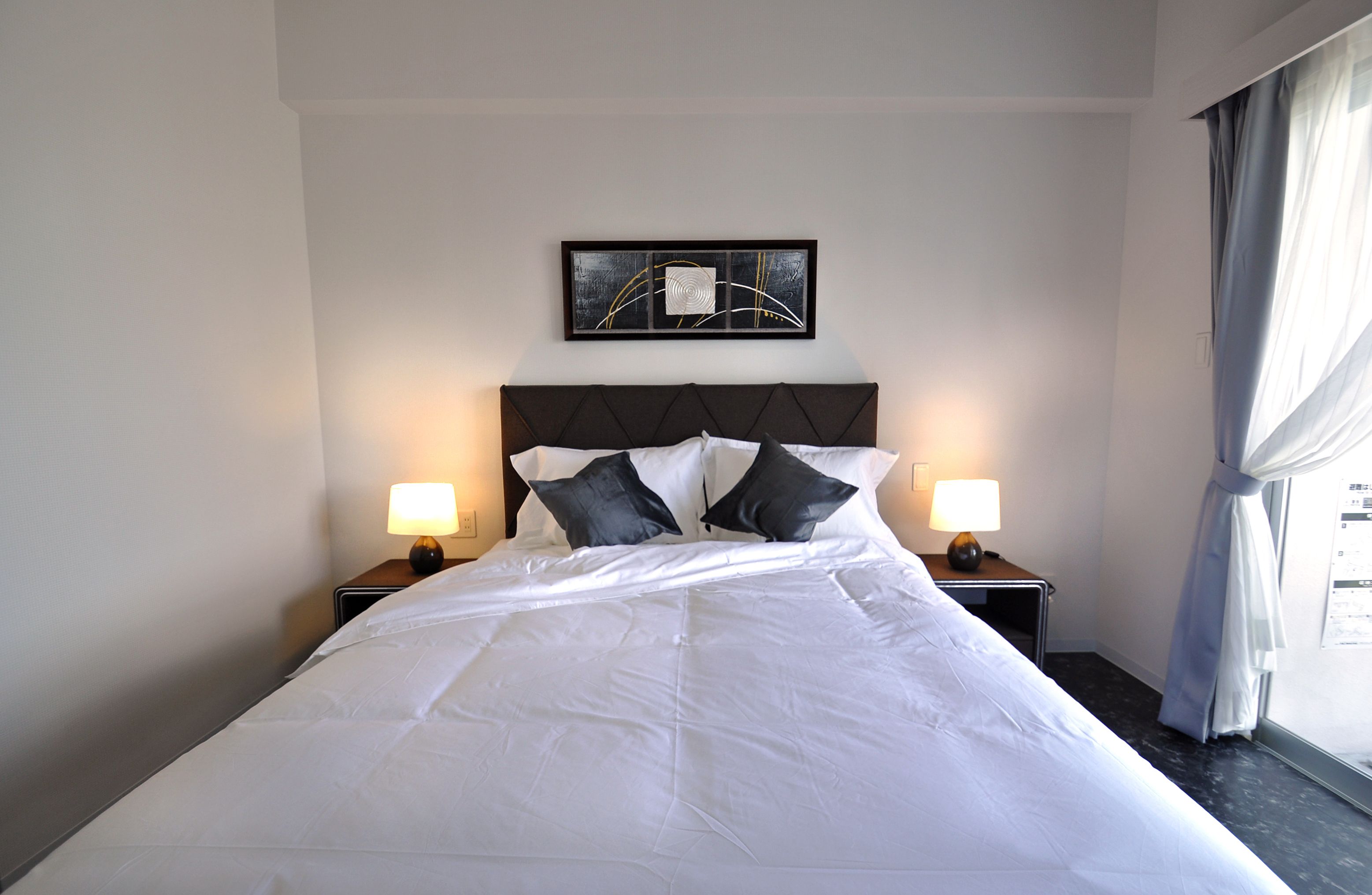 Una cama bien hecha con sábanas blancas y dos almohadas negras, flanqueada por dos mesitas de noche con lámparas, en una habitación con cabecera oscura y una obra de arte abstracta enmarcada sobre la cama.