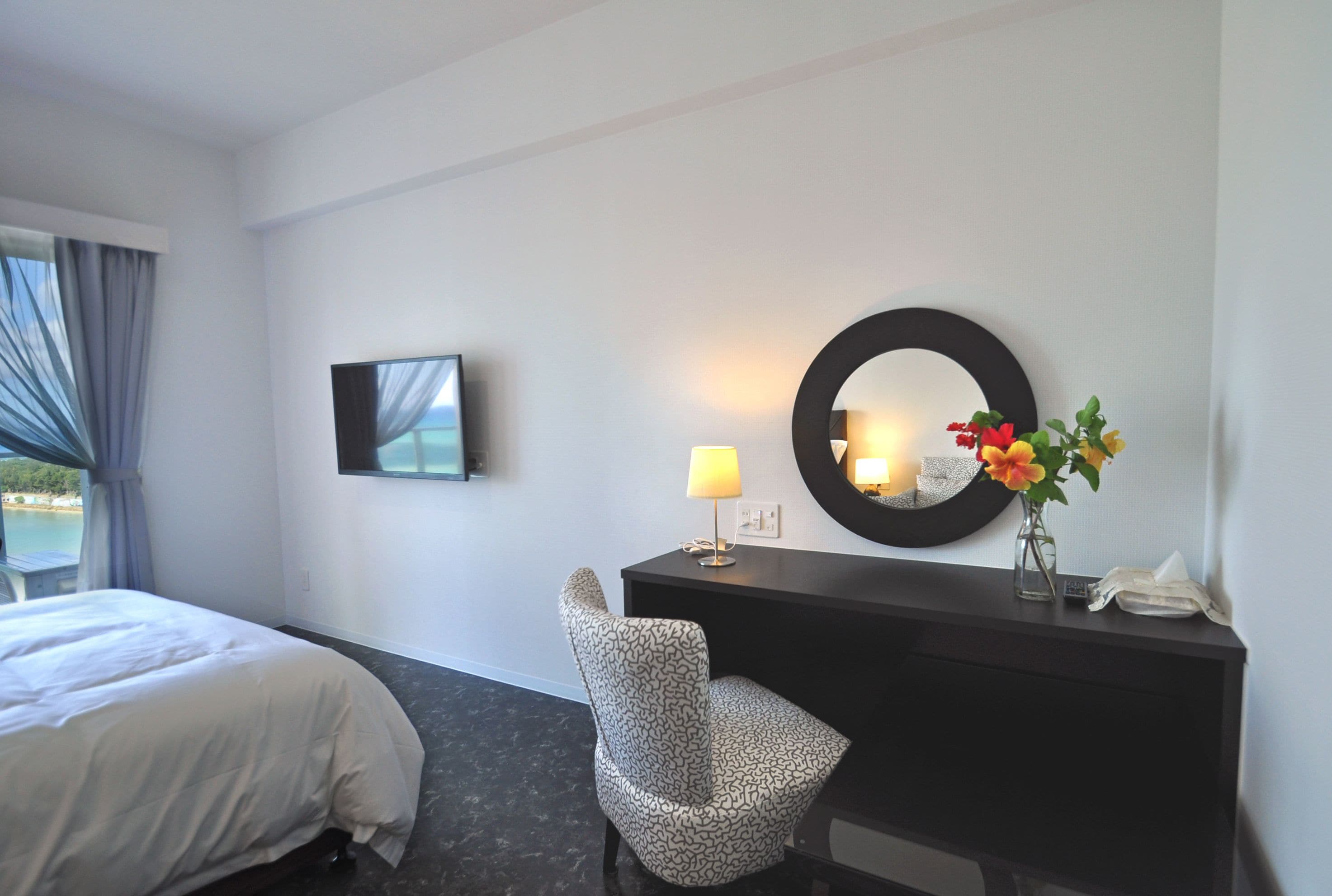 Habitación de hotel con una cama, un escritorio con lámpara, una silla, un espejo redondo y un televisor en la pared.
