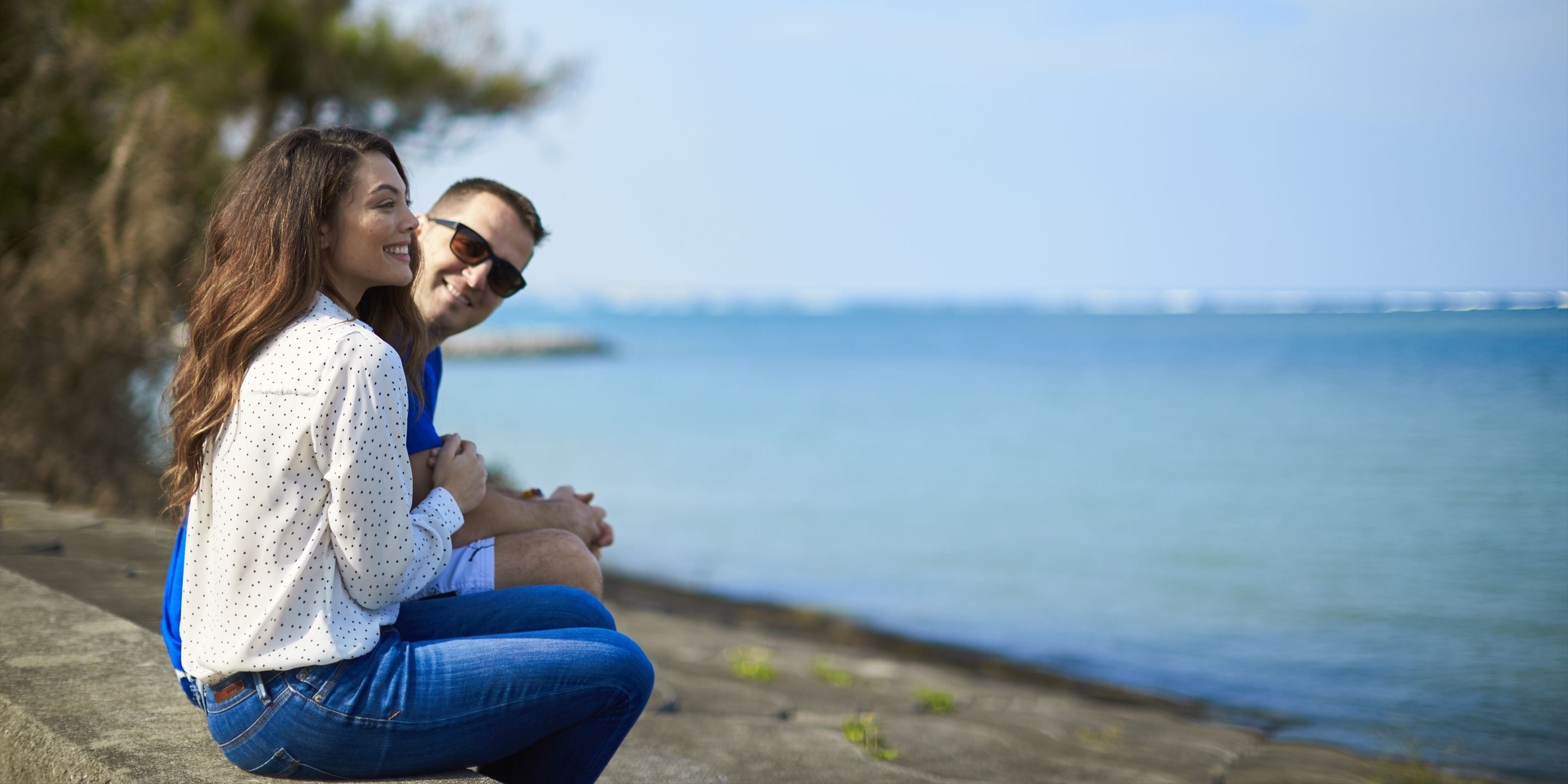 A couple sitting by a waterfront, overlooking a body of water with a distant view of a bridge.