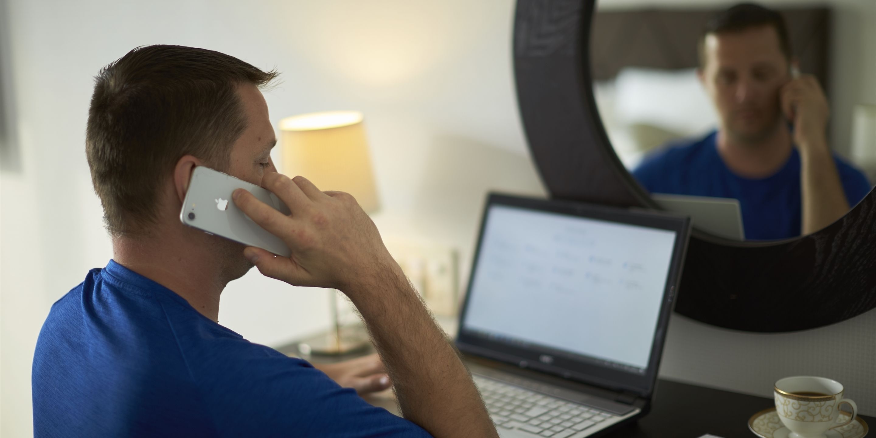 Una persona con una camisa azul usando un teléfono inteligente en un escritorio con una computadora portátil y una taza de café.