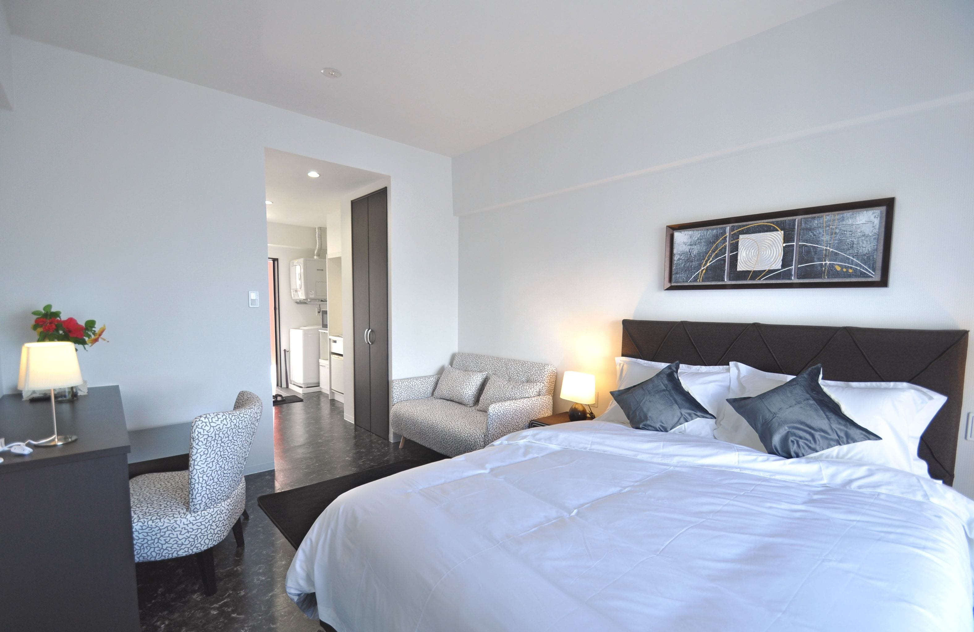 Una habitación de hotel moderna con una cama grande, una zona de estar con una silla, un escritorio y una obra de arte enmarcada en la pared.
