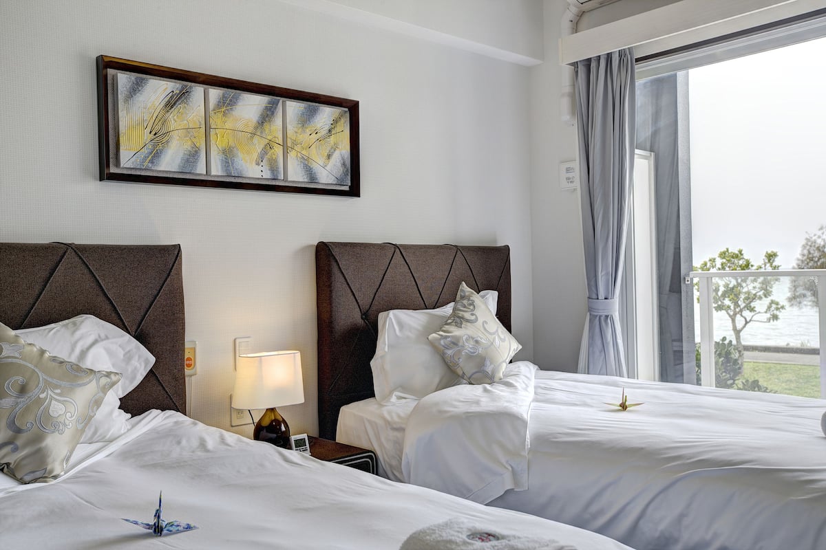 Une chambre d’hôtel avec deux lits, une table de chevet avec une lampe, un tableau abstrait encadré et une vue sur l’extérieur.