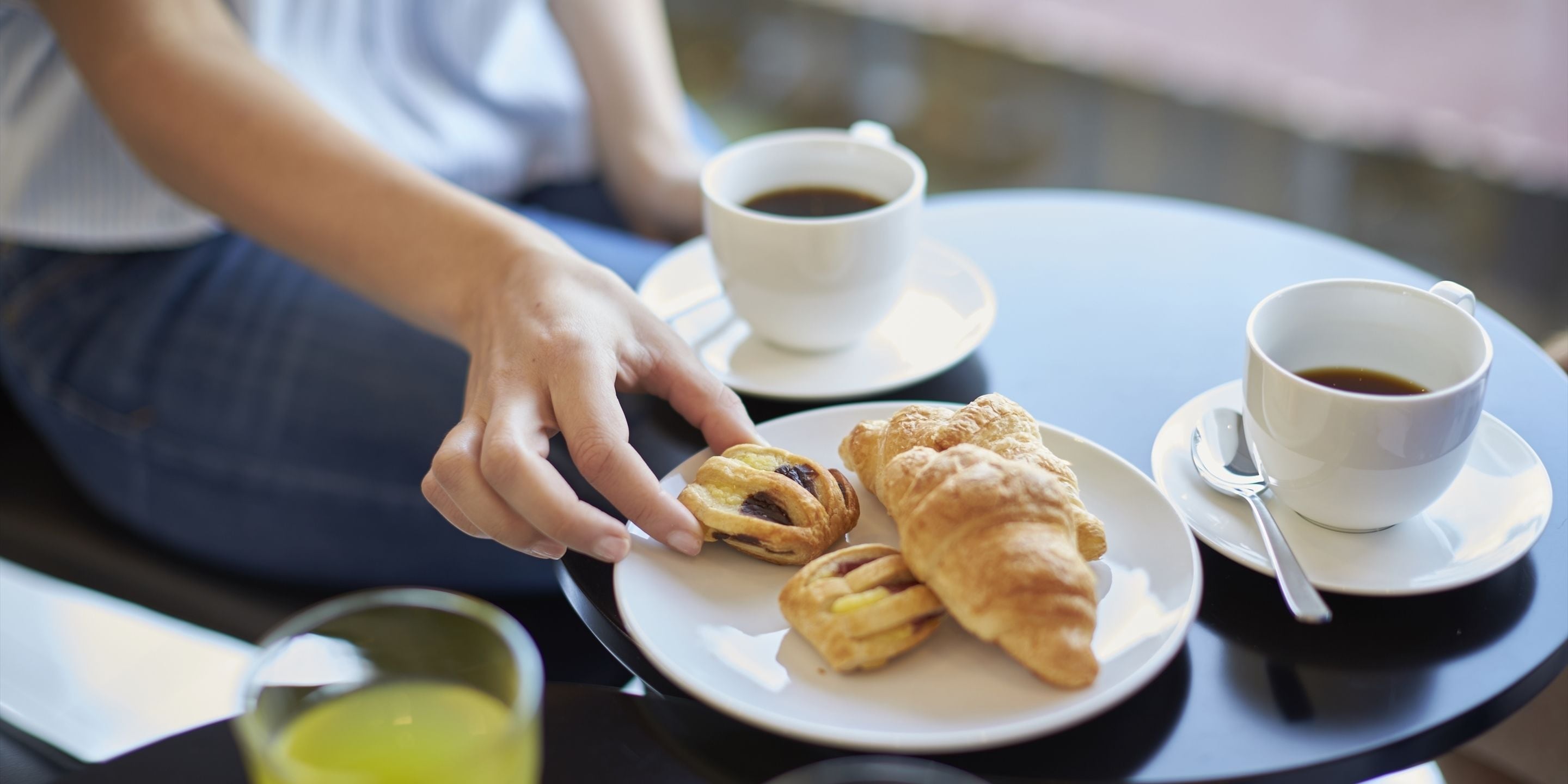 A person sitting at a table with two cups of coffee, a plate with pastries, and a glass of juice.