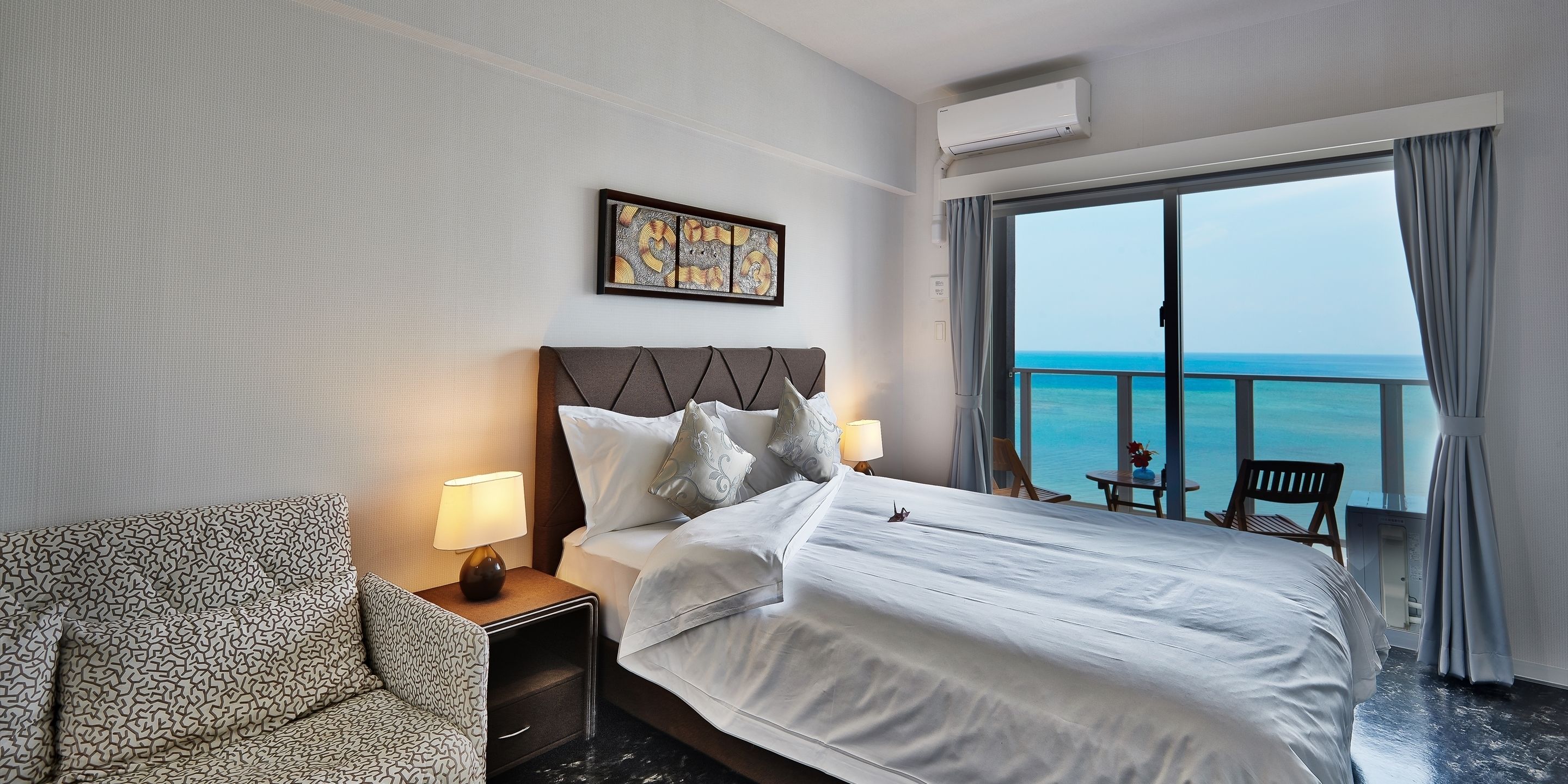 A hotel room with a large bed, a chair, a nightstand with lamps, a framed artwork, and a view of the ocean through a large window.