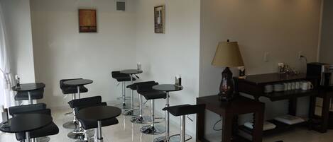 A modern hotel room with multiple black bar stools arranged around a central table, a coffee machine, and a scale on the right side.