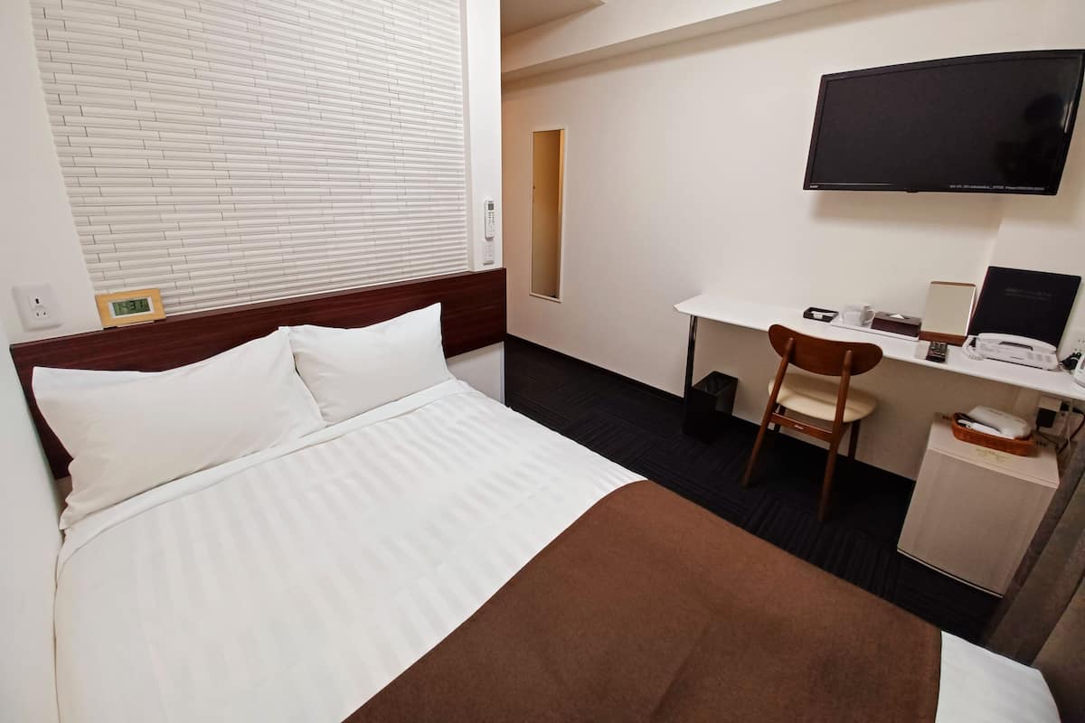 A hotel room with a bed, a desk, a chair, a TV, and a small table with a lamp.