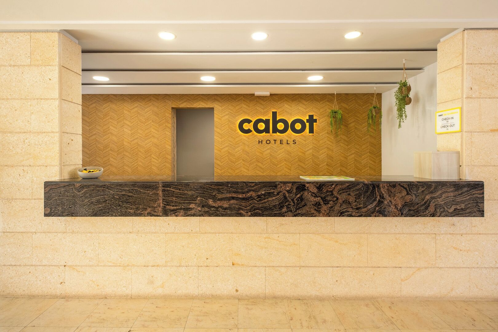 Un taulell de recepció amb una paret amb un disseny d'escama de peix, un cartell que diu 'cabot hotels' i un senyal de registre d'entrada/sortida.