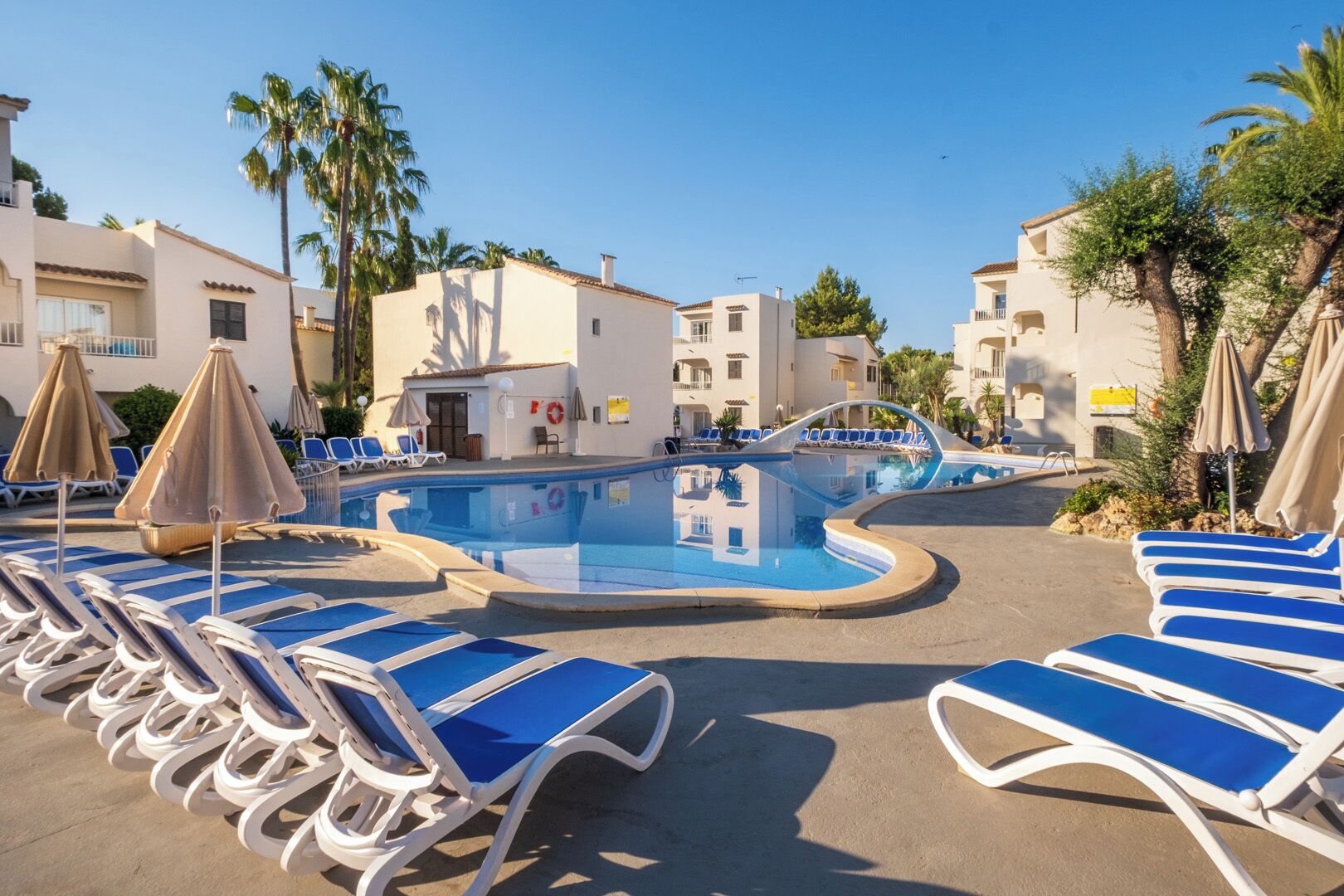 Zona de piscina con sillones de descanso y sombrillas, rodeada de edificios con balcones.