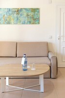 Una habitación con un sofá color beige, una mesa de centro de madera con una botella y vasos, y un cuadro abstracto de colores en la pared.