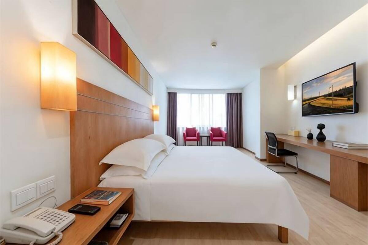 Ein Hotelzimmer mit Bett, Nachttisch mit Telefon, Schreibtisch mit Stuhl, einem Bild an der Wand und Blick ins Freie durch das Fenster.