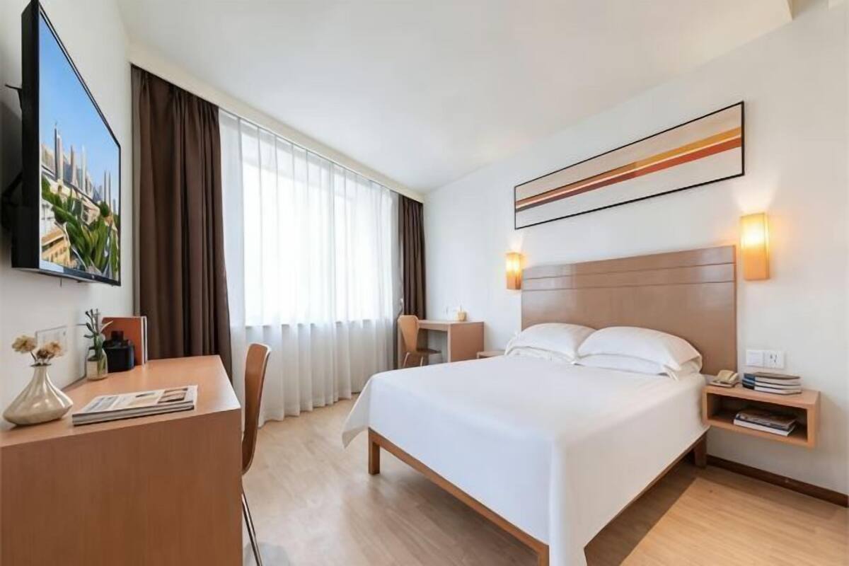 Ein Hotelzimmer mit Bett, Nachttisch, Schreibtisch, Stuhl und einem an der Wand montierten Fernseher.