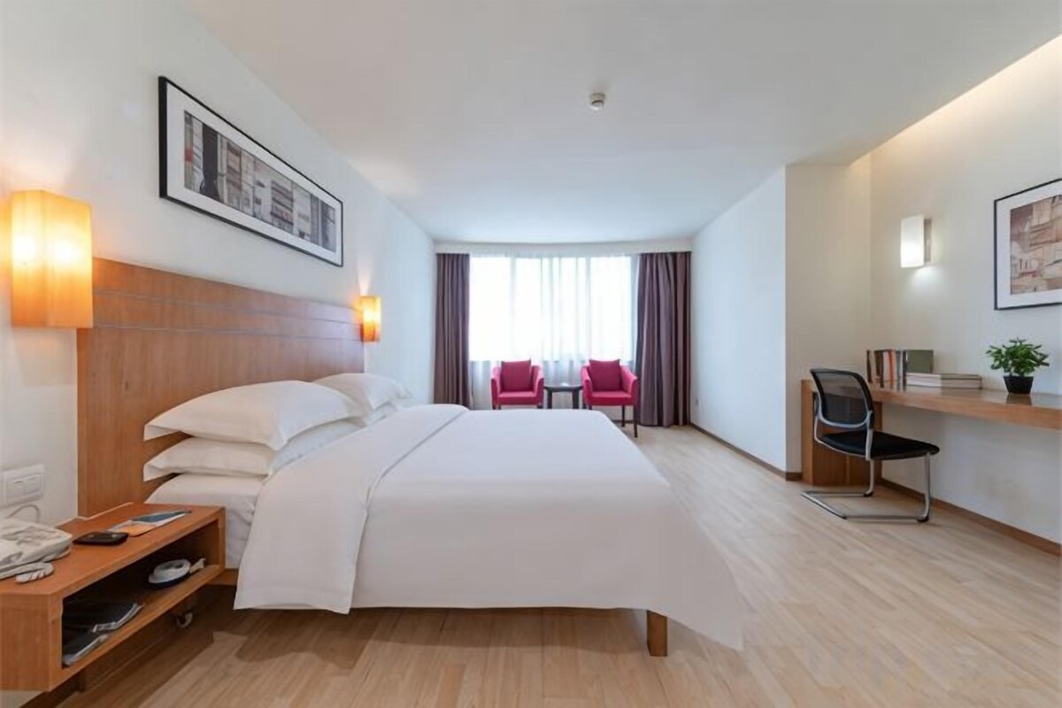 A hotel room with a bed, bedside table, lamp, and a view of the city.