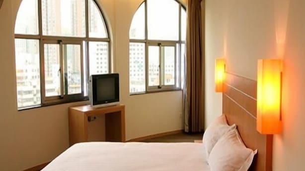 A hotel room with a bed, a television on a wooden stand, and large windows with curtains.