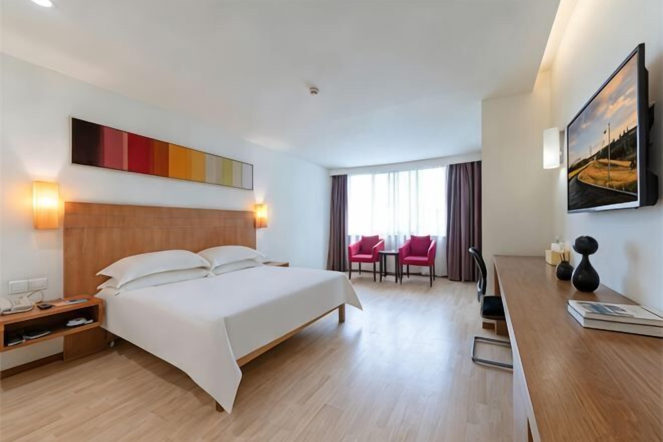 Ein Hotelzimmer mit Bett, Nachttisch, Lampe und Blick auf eine Stadtlandschaft durch das Fenster.