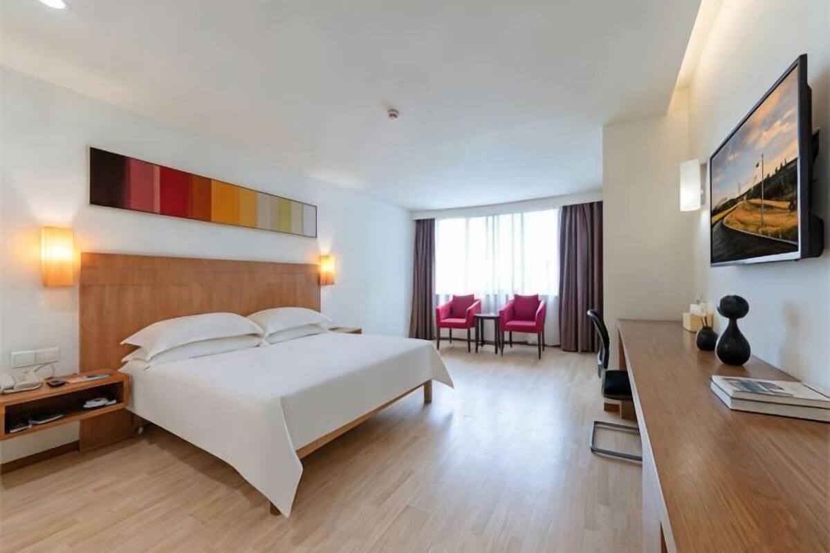 Ein Hotelzimmer mit Bett, Nachttisch, Lampe und Blick auf eine Stadtlandschaft durch das Fenster.