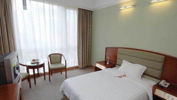 A hotel room with a large bed, a bedside table with a lamp, a chair, a small table with a vase, and a television. - Ausotel New Tianhe Guangzhou (Guangzhou)