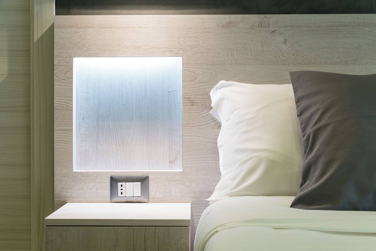 A neatly made bed with white and dark pillows, a bedside table with a switch, and a wall-mounted light fixture.