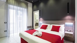 A modern hotel room with a large bed, red and white bedding, a neatly made bed with towels, a bedside table with a lamp, and a view of the balcony through sheer curtains. - Hotel Bella Napoli (Naples)